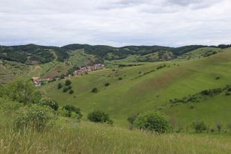 Blick über eine hochgelegene Wiese auf zahlreiche grüne Hänge im Mittelgrund sowie bewaldete Berge im Hintergrund. Links liegt eine Ortschaft zwischen den hier terrassierten Hängen.
