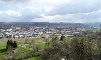 Unter einem Gewitterhimmel breiten sich bewaldete Bergketten sowie eine größere Stadt aus. Im Vordergrund Wiesen und eine von Bäumen gesäumte, kurvige Straße.