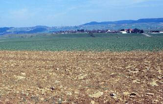 Über eine steinbedeckte Ackerfläche im Vordergrund geht der Blick zu stufigem, immer weiter ansteigendem Gelände bis zu bewaldeten Höhen im Hintergrund.