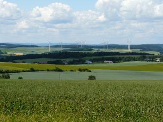 Das Bild zeigt mehrere, im Mittelgrund in einer Mulde liegende begrünte Ackerflächen. Im Hintergrund ansteigende, bewaldete Hänge lassen Platz für Windkrafträder.