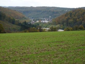 Blick über einen hochgelegen, begrünten Acker auf ein von bewaldeten Hügeln umschlossenes, besiedeltes Tal. 