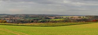 Unter einem grauen Wolkenhimmel breitet sich eine flachhügelige, von Waldgebieten durchzogene Ackerlandschaft aus. 