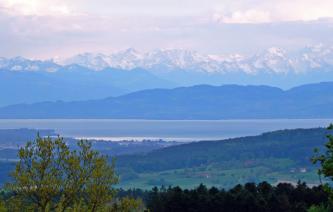 Blick von erhöhtem Standort über waldige Hügel auf einen großen See. Dahinter erheben sich bewaldete Berge sowie eine Hochbegirgskette mit verschneiten Spitzen.
