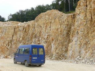 Blick von schräg rechts auf eine Abbauwand aus gelblich-beigem, eng-geklüftetem und gebanktem Gestein. Vor der Wand steht ein blauer Van.