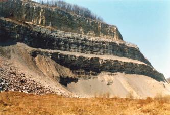 Blick von der Seite auf eine alte, schön gestufte Abbauwand eines Steinbruchs. Das anstehende Gestein zeigt eine deutliche Schichtung. Unter den drei Stufen befindet sich eine Schräge aus Geröll.