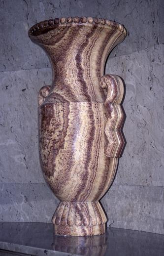 Blick auf ein steinernes Gefäß mit Sockel, trichterförmiger Öffnung oben und seitlichen Haltegriffen. Das Gestein ist gelblich bis rötlich mit dunklen, senkrecht verlaufenden Streifen.