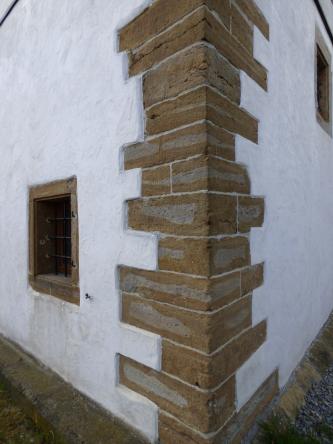 Das Foto zeigt die aus rötlich grauen Mauersteinen gefertigte Ecke eines Hauses. Die nach links und rechts abgehenden Wände des Gebäudes sind weiß.