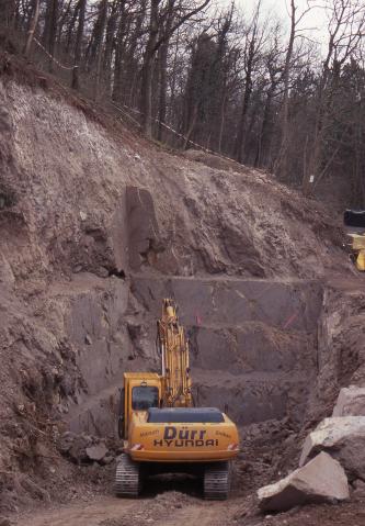 Blick auf einen nach rechts abfallenden Waldhang mit offener Seite. Das freiliegende, dunkelgraue Gestein ist in der Bildmitte wie eine Treppe zugeschnitten. Ein gelber Bagger steht davor.