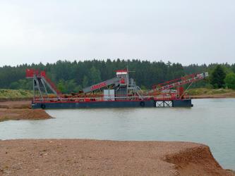 Blick auf einen See sowie einen schwimmenden Bagger mit Förderbändern. Im Vordergrund und links aufgeschüttete Ufer aus braunem Bodenmaterial. Im Hintergrund befindet sich das von Wald begrenzte Gegenufer.