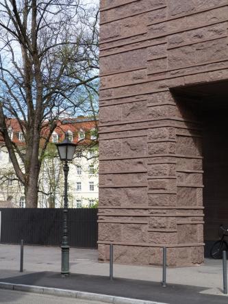 Teilansicht eines von einem rechteckigen Pfeiler gestützten Gebäudes aus braunrotem Gestein (im Bild rechts), mit Ecksteinen, dünnen Zwischenplatten und Quadern mit unterschiedlicher Oberfläche. Vor dem Gebäude verläuft ein Fußweg mit einer Laterne.