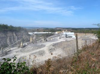 Blick über einen Drahtzaun auf ein ausgedehntes Steinbruchgelände. Auf einem Plateau, zwischen Fahrwegen und Abraumhalden, stehen Förderbänder. Links ragt eine Bruchwand auf, rechts geht es tiefer hinab. Im Hintergrund liegen bewaldete Hügel.