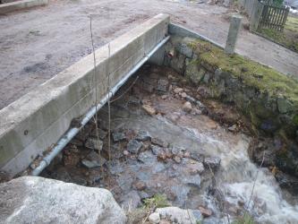 Zu sehen ist ein Teil einer steinernen Brücke, unter der ein Bach fließt. Darüber verläuft eine stark verschmutzte Straße.