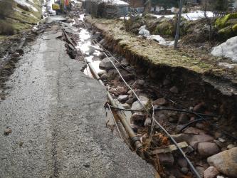 Auf dem Bild ist eine Straße, deren Asphalt auf der rechten Seite abgebrochen und so der Untergrund zu sehen ist. Zum Vorschein kommen einige Rohre und Kabel. Im Hintergrund ist ein Bagger zu sehen.